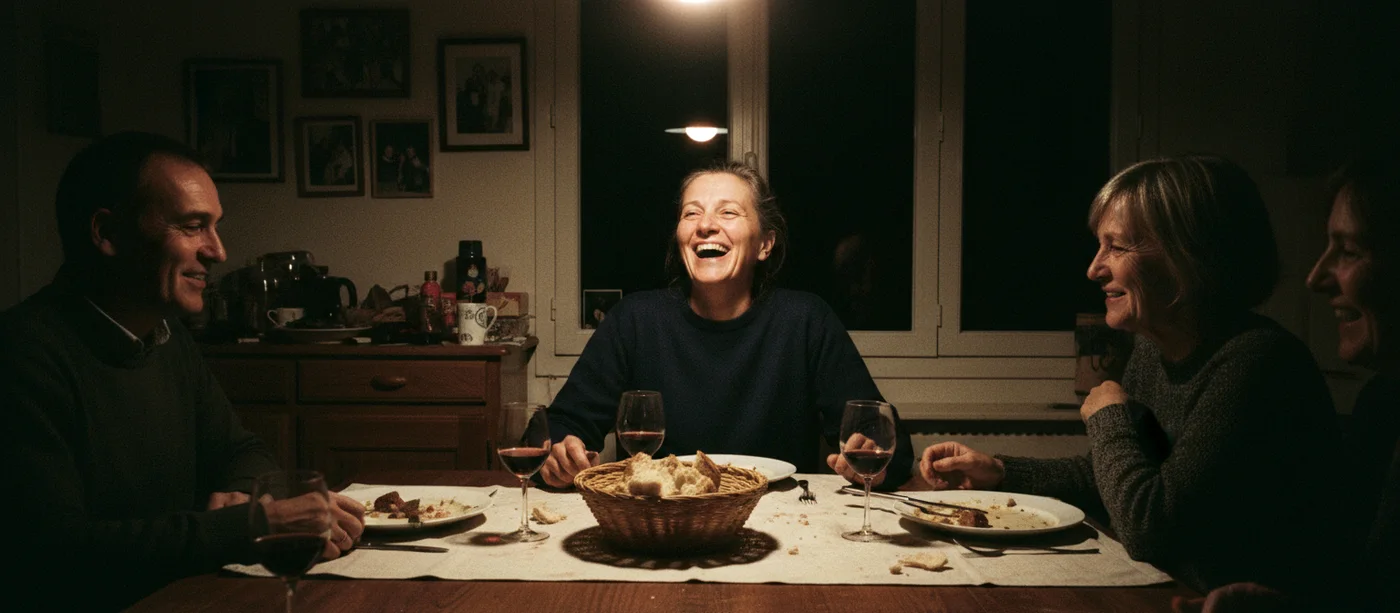 Une femme d'une cinquantaine d'années rit au milieu d'un repas de famille, le soir, dans une salle à manger française éclairée par la seule lampe du plafond, la fenêtre noire derrière elle.